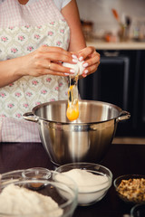 Female hands breaks a white egg into a metal bowl. Home cooking on kitchen. Plump woman in pastel apron cooks.