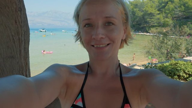 Smiling young beautiful blonde woman taking a selfie video at sandy beach by the sea. Lovrecina beach on island Brac, Croatia. Paradise on Earth. Vacation, technology a