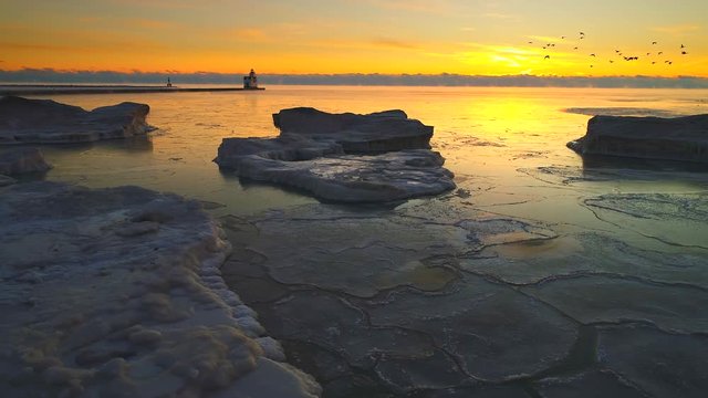 Winter Dawn Sky Over Frozen Lake Michigan Shoreline With Icebergs, Sea Smoke, Lighthouse In Distance, Moving Aerial Shot By Drone.