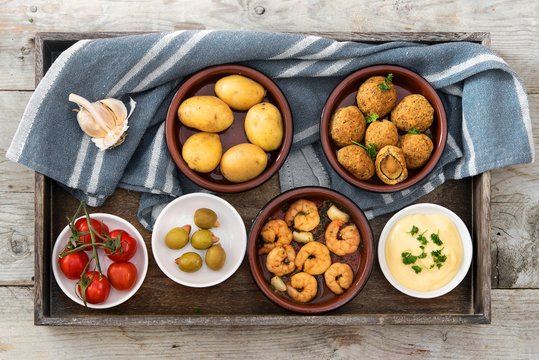 party  appetizers , spanish tapas , such as baked olives, prawn shrimps, potatoes, tomato and garlic dip on a rustic wooden tray with a blue gray kitchen towel, flat top view from above