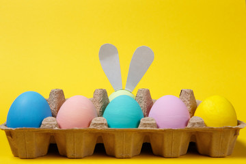 Row of colorful pastel monophonic painted Easter eggs with fun bunny ears in cardboard tray on yellow background for card. Happy Easter concept. Copy space for advertisement. With place for text.
