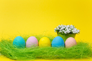 Row colorful pastel monophonic painted Easter eggs white flowers lilies of valley in bucket in green grass on yellow background. Happy Easter concept. Copy space for advertisement. With place for text