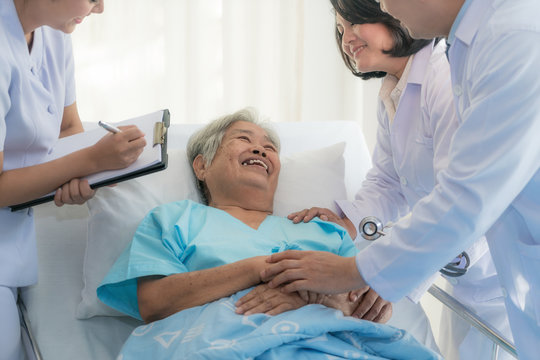 Asian Medical Team Of Doctors Examining And Talking To ASian Elderly Woman Patient, Health Care People Take Note On Clipboard In Hospital.