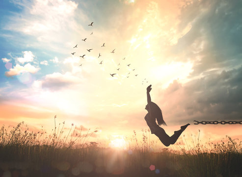 Freedom concept: Silhouette of a woman jumping and broken chains with birds flying at sunset meadow with her hands raised.