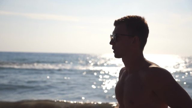 Profile of athletic guy in sunglasses jogging along sea coast. Young sporty man running on ocean beach on a sunny day. Male sportsman exercising outdoor. Healthy active lifestyle. Close up Slow motion