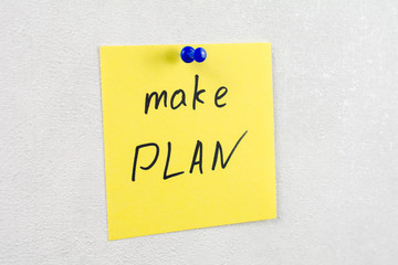 make plan note reminder yellow sticker on a white wall pinned with blue pushpin, close up, selective focus