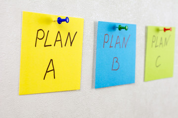 plan a plan b plan c note reminder yellow green blue sticker on a white wall pinned with blue green red pushpin, close up, selective focus