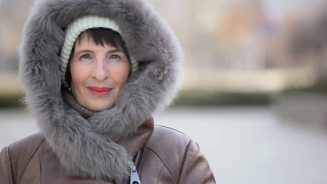 An optimistic view of a smiling brunette woman in a white knitted hat and a leather coat with a fur hood standing on winter street 