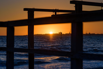 The sun sets over the horizon of Port Phillip Bay with a silhouetted wooden boat launch structure...