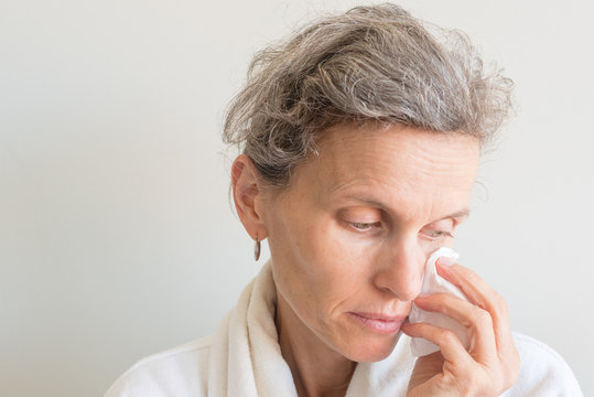 Close Up View Of Middle Aged Woman With Grey Hair In White Bathrobe Wiping Face With Tissue Against Neutral Background (selective Focus)