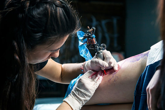 A close-up of the process of creating a tattoo of a bird with red dyes using a tattoo machine by a female tattoo master on a hip in sterile conditions in a dark workshop