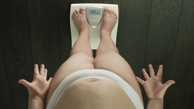 Female with extra weight looking down at scales screen, dissatisfied, body fat