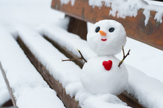 Happy snowman with a red heart on a snowy bench