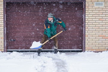 A man cleans snow in winter weather with a shovel near the garage 
