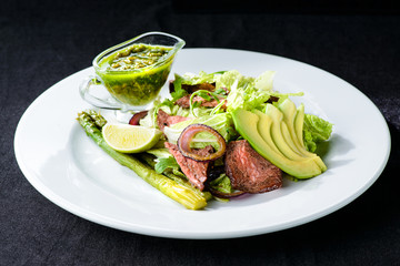 Delicious green meat salad with roasted juicy steak, avocado and green sauce in a white plate isolated on black background