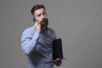 Tired overworked man on the phone holding laptop and tablet computer looking away against gray studio background. 