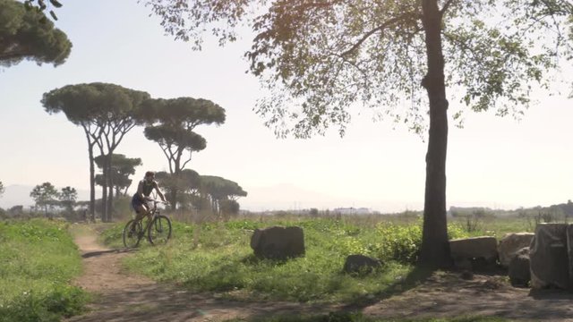 Cyclist riding bike on dirt road in in front of ancient roman aqueduct. Young attractive athletic man with blue t-shirt and shorts sportswear in parco degli acquedotti in rome