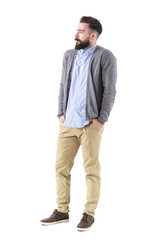 Bearded male fashion model in gray cardigan with hands in pockets looking away. Full body length portrait isolated on white studio background. 