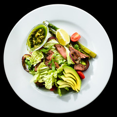 Delicious green meat salad with roasted juicy steak, avocado and green sauce in a white plate isolated on black background. Top view, flat lay