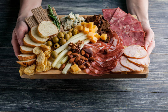 Female Hands Hold Assorted From Meat And Cheese On A Chopping Board. Smoked Sausage, Meat Roll From A Turkey With Cheese, Jamon, Olives, Nuts And Various Grades Of Cheese. Soft Focus. Low Key.