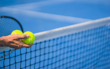 old asian man hold two tennis balls in right hand, selective focus, blurred racket, net and blue tennis court as background, “aging population” concept