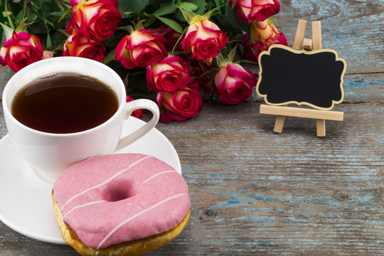 Cup of tea with donut, fresh roses and blackboard with space for your text  on a wooden background. Perfect image for mother's day