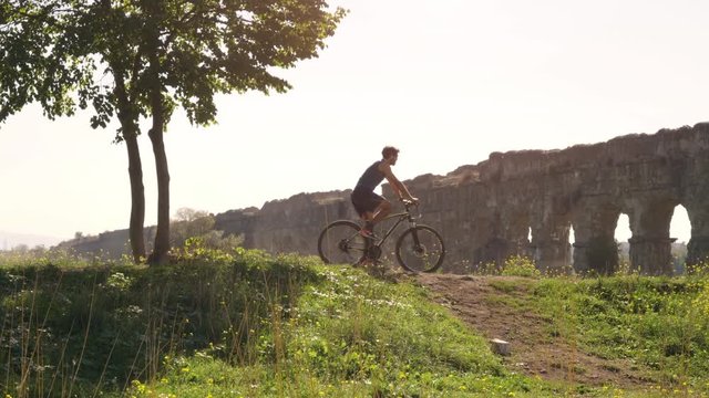 Cyclist riding bike uphill on dirt road in front of majestic ancient roman aqueduct. Young attractive athletic man with blue t-shirt and shorts sportswear in parco degli acquedotti in rome