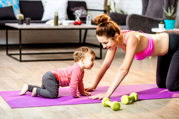 Young mother stretching on the mat with her baby son playing on the floor at home