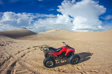 Red quad vehicle on sandy dune. Blue sky over desert landscape.