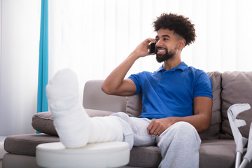 Happy Young Man With Broken Leg Talking On Mobile Phone