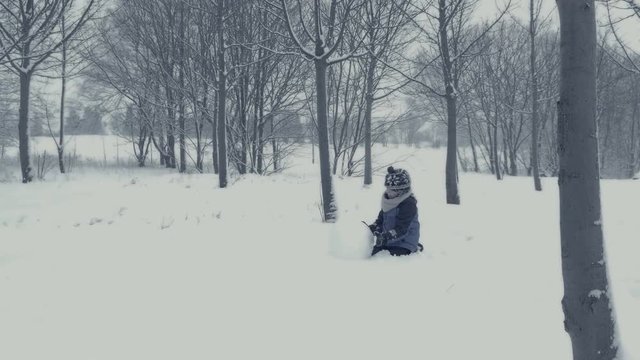 Small boy builds snowman. Family plays in snow. Yougster enjoys winter fun. Winter landscape with child and falling snow. Everything is covered with fresh powder. Winter background, Christmas theme.
