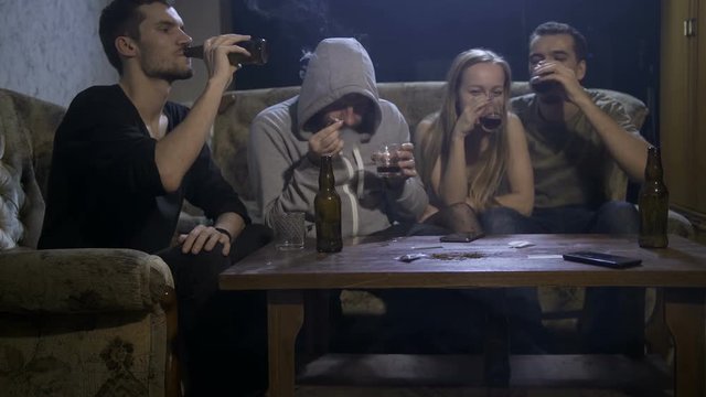 Four young addicted people sitting in domestic interior on the sofa at night, drinking alcohol while man in hooded shirt smoking cannabis joint. Social issues, alcohol and drug abuse concept