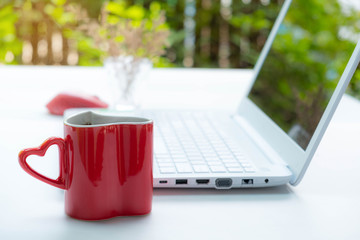 Heart shape red cup of coffee with steam on top , there are white notebook computer in the background on white table in the garden