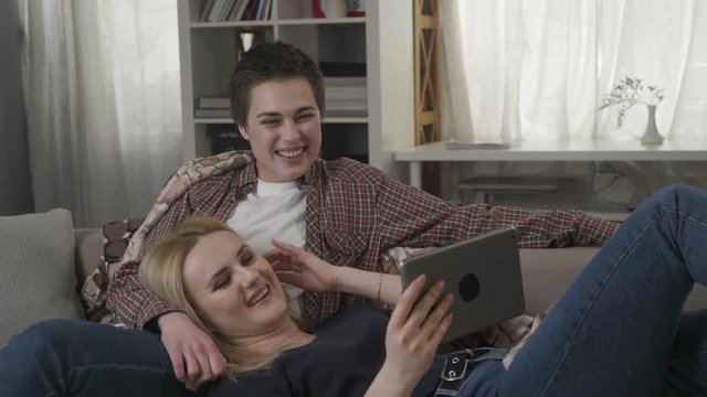 Lesbian couple is resting on the couch, using tablet and laughing, young family, plaid, cozy 60 fps