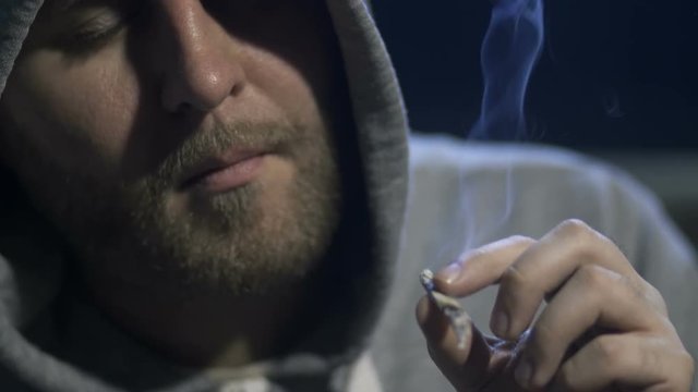 Close-up young male portrait smoking marijuana joint at home in a domestic room at night. Addicted man in a hooded shirt inhaling cannabis smoke. Social issues and drug addiction concept