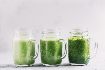 Variety of three color green spinach kale apple yogurt smoothie in mason jars in row over gray background. Healthy vegan detox eating. Toned image