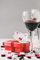 Love Valentine day greeting card with two glasses of red wine, red gift box with orchid flower and wooden hearts over white gray background. Copy space. Toned image