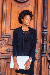 Young African American Businesswoman with afro hairstyle working in New York, wearing black fashionable clothes, wristwatch, holding laptop computer, standing by vintage office doorway, looking away..