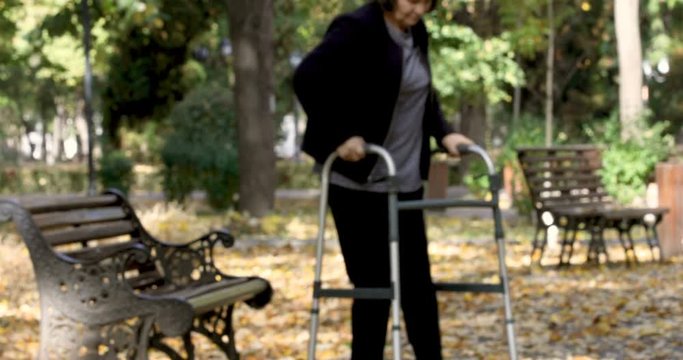 Senior woman with walker getting up from bench and walking outdoors in autumn park. The person comes in focus.