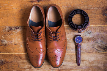 Closeup of elegant stylish brown male formal accessories isolated on brown wooden background. Top view of leather belt, shoes and watch. Preparation for wedding or bussiness day concept. 