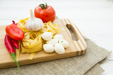 Healthy Food, Kitchen Concept. A set of cooking ingredients on a wooden cutting board: fetuccini nests, tomato, chilli peppers, garlic and mozarella cheese, white wooden background
