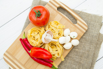 Healthy Food, Kitchen Concept. A set of cooking ingredients on a wooden cutting board: fetuccini nests, tomato, chilli peppers, garlic and mozarella cheese, white wooden background, top view