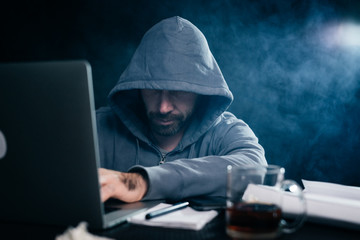 A computer hacker is typing on a laptop in a smoky room. Dark background