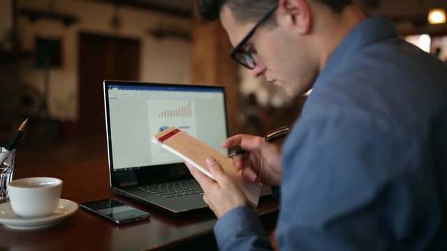 Hurried handsome young male businessman in glasses working on laptop with diagrams on sreen, checking time on smartphone app and taking notes in notepad. Multitasking telecommuting concept.