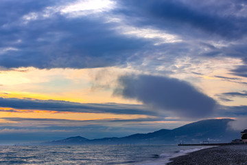 High clouds and fog on the coast, the sun is hidden behind the clouds, on the shoreline are visible the houses of the city on the hills. Beautiful colorful sunset on the Black Sea, Sochi, Russia.