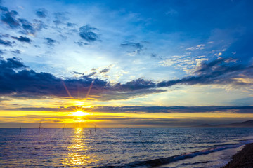 The bright rays of the yellow sun illuminates the sky with thick blue clouds and the evening sea on the shore. Beautiful colorful sunset on the Black Sea, Sochi, Russia.