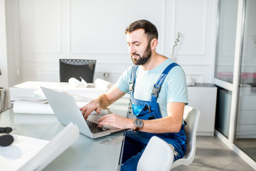 Handsome repairman or foreman in uniform working with laptop and architectural drawings at the...