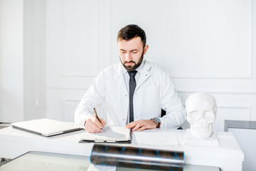 Fototapeta premium Handsome male doctor in the medical gown writing scientific works sitting in the beautiful white office interior