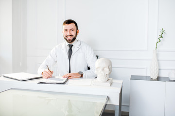 Portrait of a handsome male doctor in the medical gown writing scientific works sitting in the beautiful white office interior