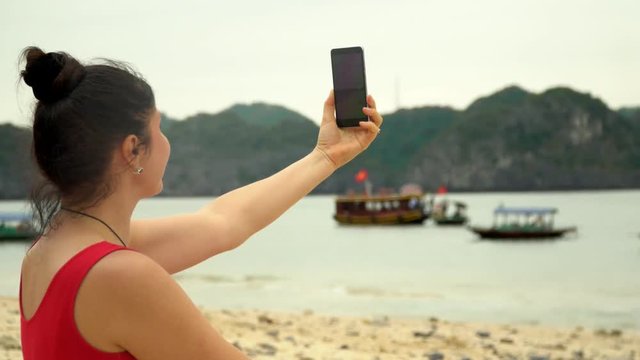 Beautiful woman using smart phone on beach. The traveler on the island of monkeys. The woman takes the picture against the background of a bay with boats.